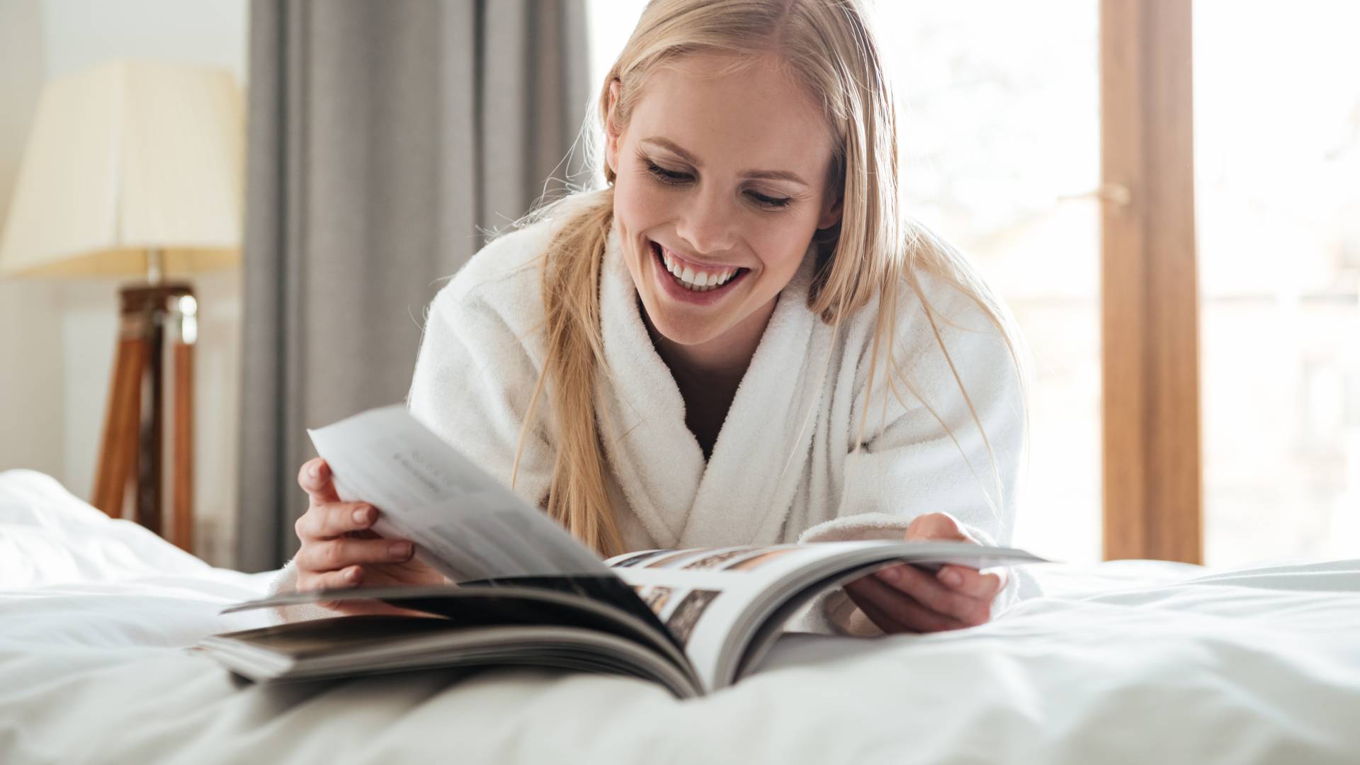 Frau liegt mit Morgenmantel im Bett und ließt eine Zeitschrift