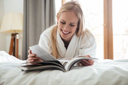 Frau liegt mit Morgenmantel im Bett und ließt eine Zeitschrift